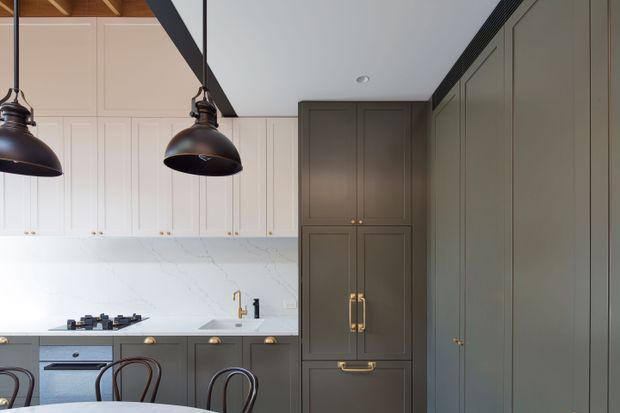 A refined kitchen interior featuring full-height dark grey-green cabinetry, pale shaker-style wall cabinetry, a white marble-look splashback and benchtop, brass hardware and tapware, and oversized dark pendant lights. Timber ceiling framing and dining chairs are visible, creating a warm, sophisticated contrast.