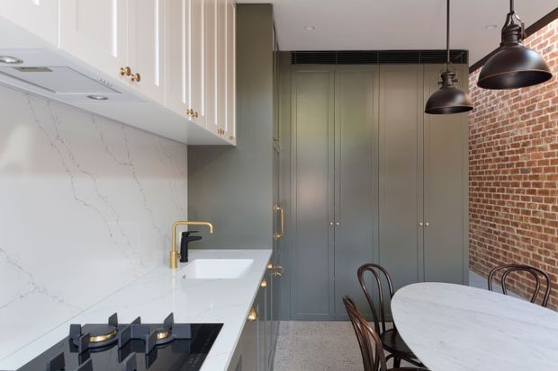 A compact contemporary kitchen with white upper cabinetry, a marble-look splashback and benchtop, black gas cooktop, and a gold-and-black tap. Tall muted grey built-in cabinetry lines the rear wall, while a rounded white dining table and bentwood chairs sit beside an exposed red brick wall. Black industrial pendant lights hang overhead.