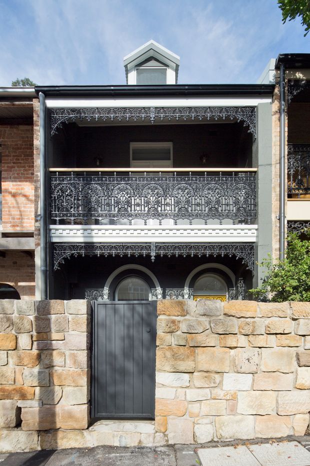 Front exterior view of a narrow two-storey heritage terrace house with elaborate cast-iron lacework verandahs and balcony balustrades, arched ground-floor windows, a projecting dormer, and a sandstone boundary wall with a dark metal pedestrian gate. The facade combines exposed red brick, painted masonry, and intricate ornamental metalwork in a dense historic streetscape.