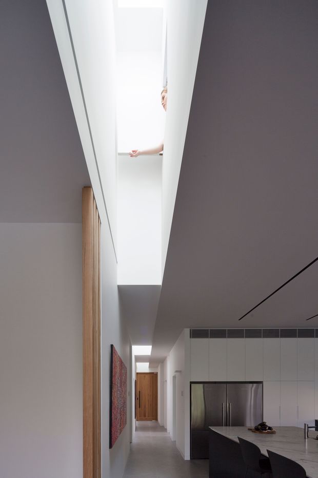 A bright, minimalist contemporary interior showing a long hallway opening into a kitchen. The space features white walls and cabinetry, a pale stone kitchen island, dark cabinetry accents, integrated stainless-steel appliances, timber doors and vertical timber detailing, recessed linear lighting, and a dramatic elongated skylight or clerestory bringing daylight into the corridor. A colorful artwork adds a small accent along the hallway.