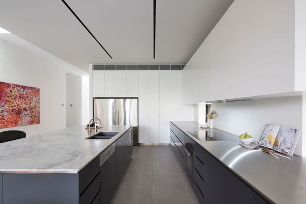 Contemporary galley-style kitchen with a large waterfall-edge stone island, dark built-in cabinetry, stainless-steel worktops and appliances, and white full-height cabinetry. Linear recessed ceiling details, minimal hardware, and integrated shelving create a clean, refined interior. An adjacent dining or living area is partially visible.