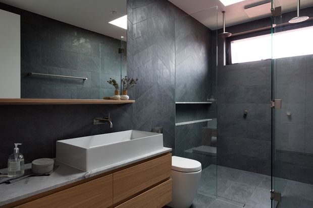 Contemporary bathroom with dark grey stone-look wall and floor tiles, a white vessel basin on a timber vanity, open recessed shelving, toilet, and a frameless glass shower enclosure. The space has restrained dark finishes, warm timber joinery, and a skylight or high window providing natural light.
