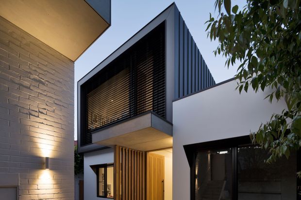 Contemporary multi-level residential facade viewed from an outdoor approach, featuring crisp white rendered walls, dark metal vertical cladding, a projecting upper volume with louvred screening, warm timber entry detailing, and exposed brickwork at the left. Integrated wall lighting and mature planting frame the entrance.