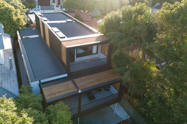 Aerial view of a contemporary multi-level residential home nestled among mature trees. The building features dark corrugated metal roofing, warm timber-clad wall sections, extensive outdoor timber decking, covered balconies, and strong horizontal roof and deck lines. Surrounding neighbouring homes and dense greenery are visible.