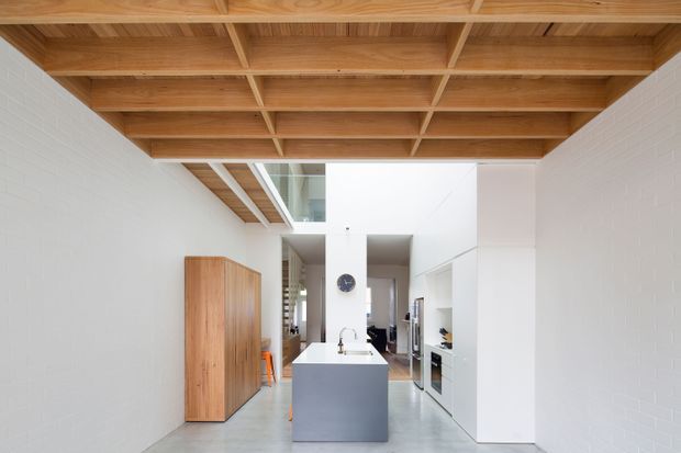A bright, narrow contemporary kitchen and living circulation space with a large central island, white painted brick walls, polished concrete flooring, extensive white built-in cabinetry, and a striking exposed timber ceiling grid. Natural light enters through high-level glazing and a roof opening, while a timber storage unit adds warmth.