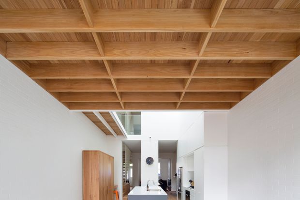 Bright contemporary interior with a dramatic exposed timber ceiling featuring a regular coffered beam grid. White painted brick walls and white built-in cabinetry surround a compact kitchen area, with a timber storage cabinet and a partial view toward adjoining spaces. A glazed clerestory-like opening brings additional light into the room.