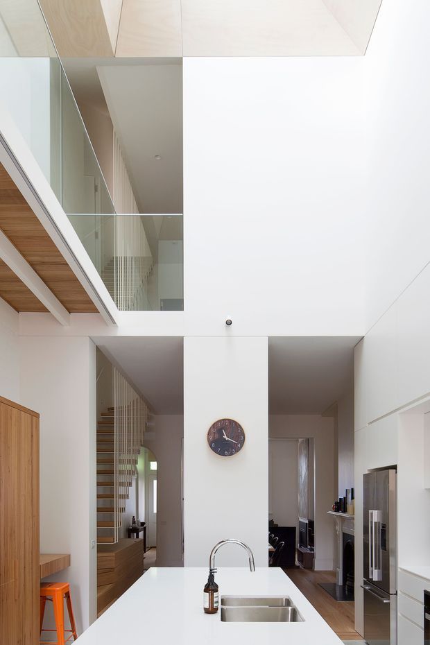 Bright contemporary open-plan kitchen viewed toward a double-height void and stairwell. The space features a large white island with an undermount stainless-steel sink and curved chrome tap, white full-height cabinetry, stainless-steel appliances, warm timber cabinetry and ceiling lining, and a glass balustrade overlooking the stairs. A dining or living area is visible beyond the kitchen.