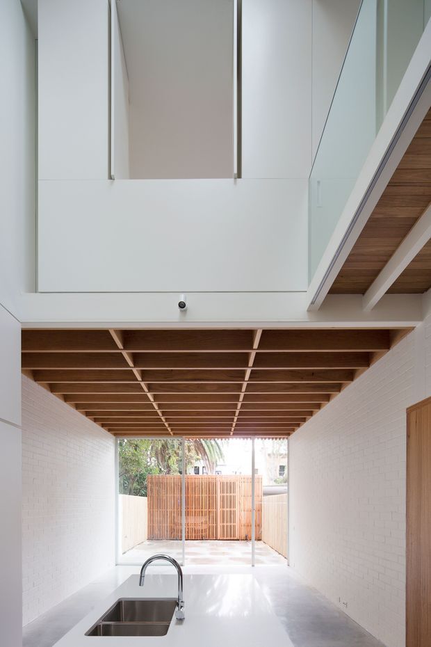 A bright, narrow contemporary kitchen or utility space beneath a timber-lined mezzanine or stair structure. White-painted brick walls and white cabinetry create a clean backdrop, with a long pale benchtop, stainless-steel sink and gooseneck tap in the foreground. Exposed timber ceiling battens and beams add warmth, while full-height glazing opens to a small timber privacy-fenced courtyard. A stair or upper-level walkway with timber soffit is visible at upper right.