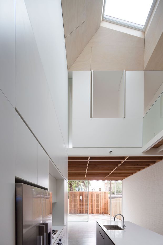 A tall, narrow contemporary kitchen opening toward an outdoor courtyard. The space features extensive white built-in cabinetry, integrated appliances, a white island with a stainless-steel sink and curved tap, pale timber wall and ceiling surfaces, a timber slatted ceiling section, and a large skylight creating a bright double-height feel.