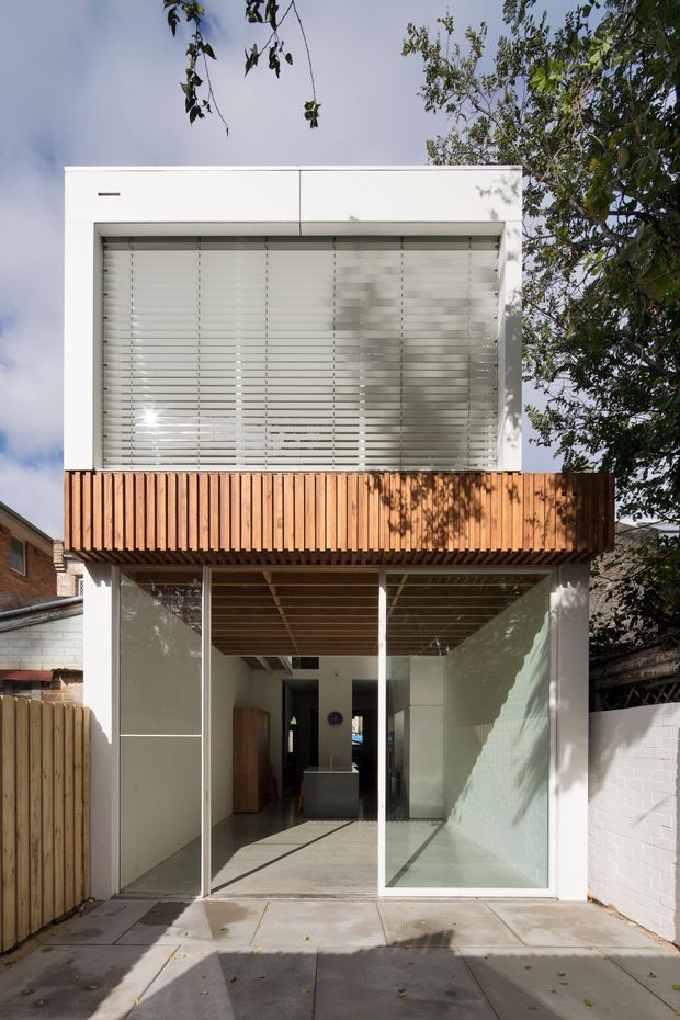 A narrow contemporary two-storey residential extension viewed from a paved rear courtyard. The white rendered facade has large full-height glazed doors or windows at ground level, a timber slatted soffit, and an upper-level balcony or screened opening enclosed by horizontal white louvres. Warm vertical timber screening forms the balcony balustrade, with neighbouring brick walls and mature trees surrounding the building.