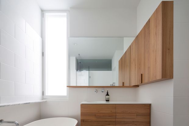 Bright contemporary bathroom featuring white tiled walls, a freestanding bathtub, timber vanity cabinetry, a long mirrored wall cabinet, and a narrow vertical window. A glass shower enclosure is visible in the mirror reflection.