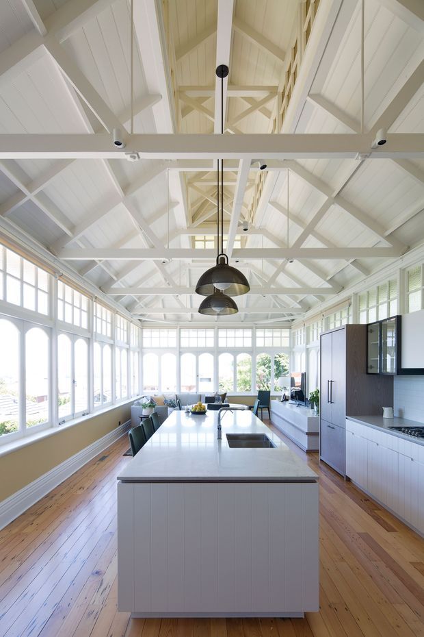 Bright, spacious kitchen in a converted or traditional timber structure, featuring a large white island with integrated sink, white cabinetry, dark pendant lights, timber flooring, and extensive floor-to-ceiling windows. The vaulted ceiling has exposed white-painted timber trusses and rafters, creating an airy, light-filled interior.