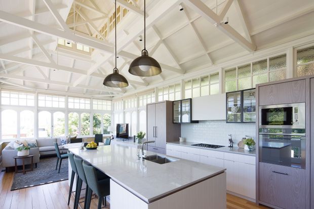 Bright, spacious open-plan kitchen and living area with a large white stone island, integrated appliances, white cabinetry, and dark pendant lights. The room features extensive gridded glazing, a high pitched ceiling with exposed white-painted trusses, timber flooring, and a small adjacent dining and sitting area.