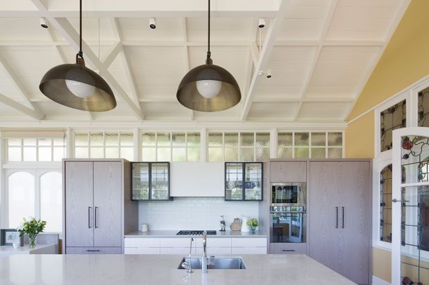 Bright, refined kitchen with a large pale stone island and integrated sink in the foreground, light grey cabinetry, a tiled backsplash, framed glass-front cabinetry, and a wide bank of high windows. The room features a white coffered or beam-detail ceiling with two oversized bronze pendant lights, warm yellow walls, and decorative stained-glass door panels at the right.