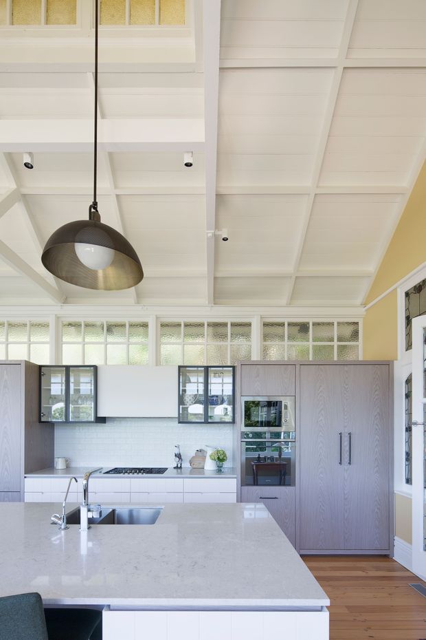 Bright, refined kitchen with a large pale stone island, white cabinetry, integrated appliances, and grey timber-grain tall joinery. The room features a high coffered white ceiling, glazed clerestory-style internal windows, warm yellow walls, timber flooring, and a large brushed-metal pendant light.
