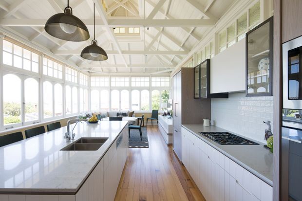 Bright, spacious kitchen in a glazed sunroom-like setting, featuring a large white stone island with integrated sink, white shaker-style cabinetry, white subway-tile splashback, timber flooring, dark-framed upper cabinetry, and exposed white-painted roof trusses. Arched windows and abundant glazing create an airy, refined atmosphere.