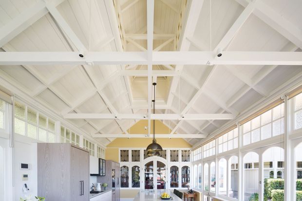 Bright, elongated open-plan interior with a high pitched ceiling and exposed white-painted timber trusses and beams. Large multi-pane windows and glazed arched openings line the sides, bringing in abundant daylight. A contemporary kitchen with tall pale cabinetry, island, pendant lighting, and glass-fronted display joinery occupies the space, with a refined light neutral palette and strong architectural detailing.