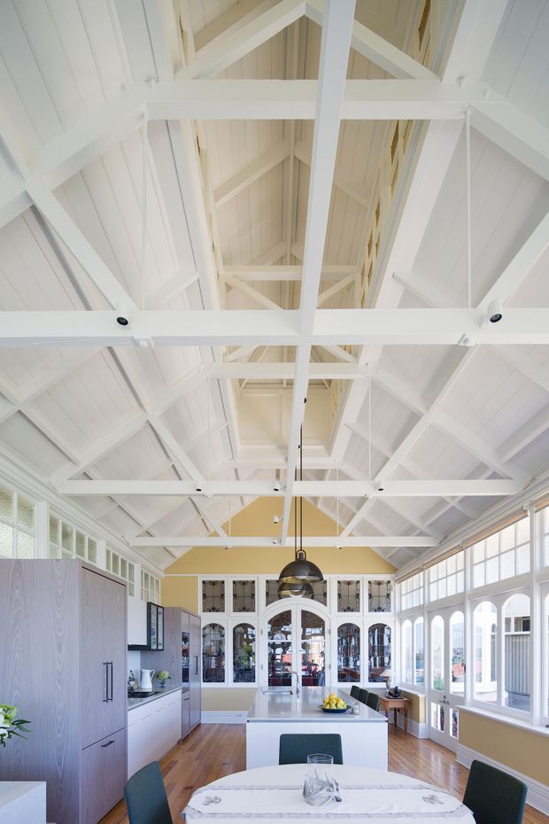 Bright, spacious kitchen and dining area beneath a dramatic white exposed timber truss roof. The room features extensive multi-pane glazing, white cabinetry, a central island with pendant lighting, warm timber flooring, pale yellow walls, and a dining table in the foreground.