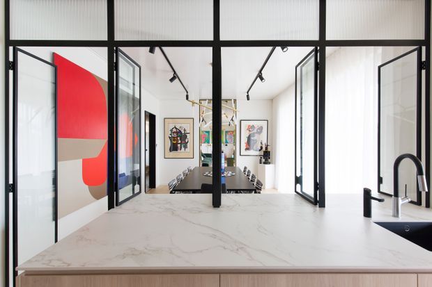 Bright contemporary kitchen viewed across a large white veined stone island, with pale timber cabinetry, matte black framed glass partitions and doors, black tapware, and a dining area beyond. A bold red and neutral abstract artwork adds colour, while black track lighting and extensive glazing create a crisp, open feel.