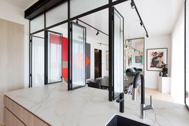 Bright contemporary kitchen with a large white veined stone island benchtop and undermount sink, viewed toward a black-framed glazed partition and adjoining living area. The space features pale timber cabinetry, black-framed internal glazing with fluted glass panels, track lighting, and colourful artwork visible beyond.