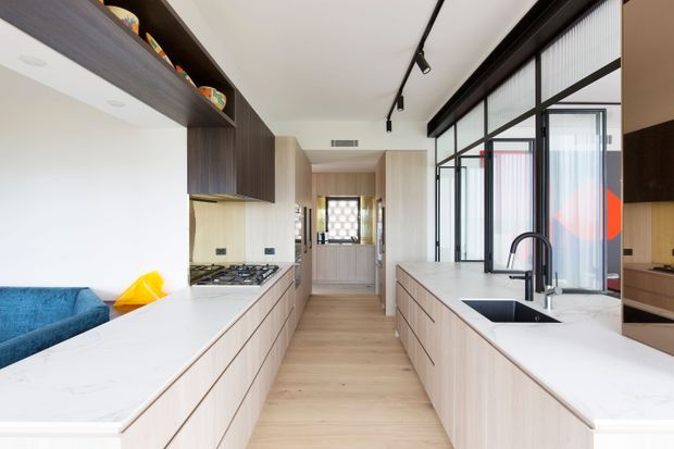 Bright contemporary open-plan kitchen with two long light-wood island runs, white stone-look benchtops, integrated cabinetry and black-framed glazed partitions. A gas cooktop, undermount sink with dark tapware, track lighting and a compact living area are visible, with a hallway extending toward the rear of the home.