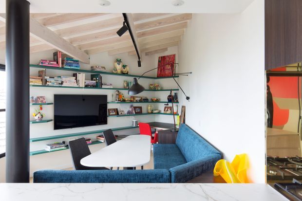 A bright, compact contemporary living and work area viewed from behind a marble-topped counter. The room features a blue built-in or sectional sofa, white sculptural table, desk chairs, extensive open shelving with books and decor, a wall-mounted television, black track lighting, exposed pale timber ceiling beams, and a dark vertical structural column. A kitchen or built-in joinery area is partially visible through the opening at right.
