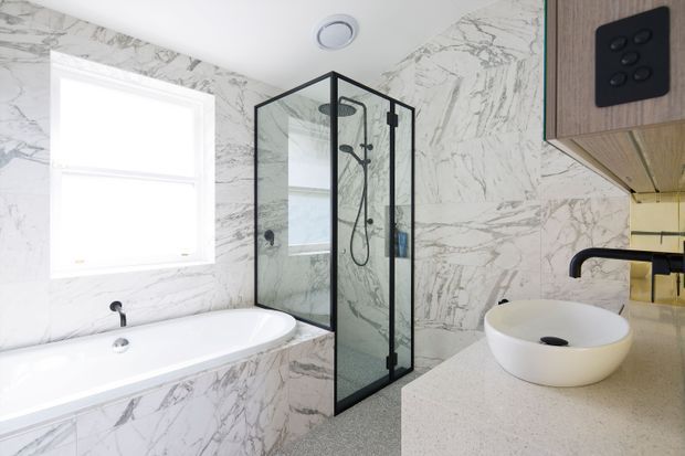 Bright contemporary bathroom featuring a freestanding-style white vessel basin on a pale benchtop, a built-in bathtub, and a black-framed glass shower enclosure. White marble-look wall and bath-panel surfaces create a clean, high-contrast finish, with timber-toned cabinetry and a black tapware and switch accents.