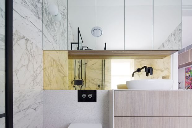 Contemporary bathroom vanity area with a freestanding white basin, matte black tapware and shower fittings, pale timber cabinetry, white marble-look wall surfaces, and a mirrored overhead cabinet. A translucent yellow-toned glass splashback adds a distinctive feature behind the basin and shower zone.