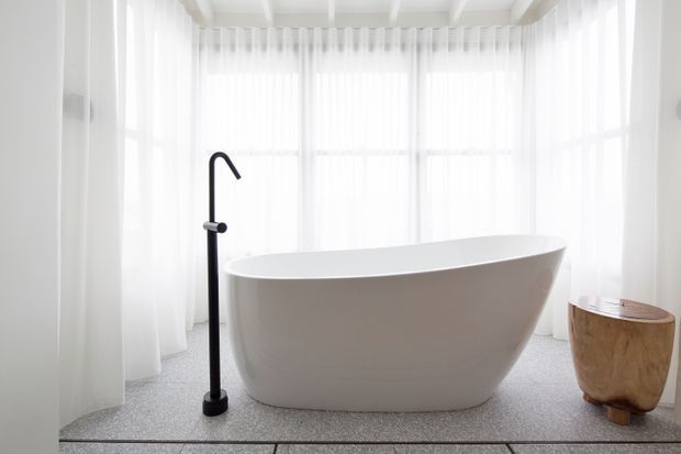 Bright, minimalist bathroom featuring a freestanding white oval bathtub positioned in front of sheer floor-to-ceiling curtains and large windows. A matte black floor-mounted bath filler stands beside the tub, with a small natural timber stool or side table nearby. The ceiling has exposed white-painted beams, and the space is softly lit with a clean, serene atmosphere.
