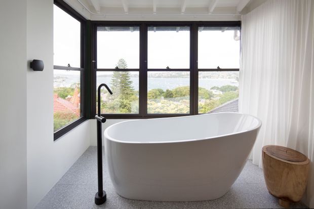 Bright contemporary bathroom featuring a freestanding white oval bathtub positioned in front of large black-framed corner windows with expansive leafy suburban views. The room has a light grey textured floor, white walls and ceiling, sheer white curtains, matte black bath fittings, and a small natural timber stool.