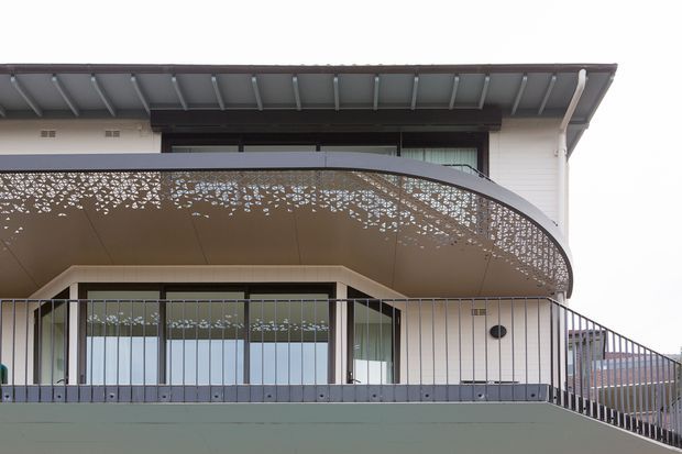 Close-up exterior view of a contemporary multi-level residence featuring a broad curved balcony canopy with intricate perforated metal screening, slim metal balustrades, light horizontal wall cladding, large glazed doors and windows, and a projecting roof with exposed structural framing.