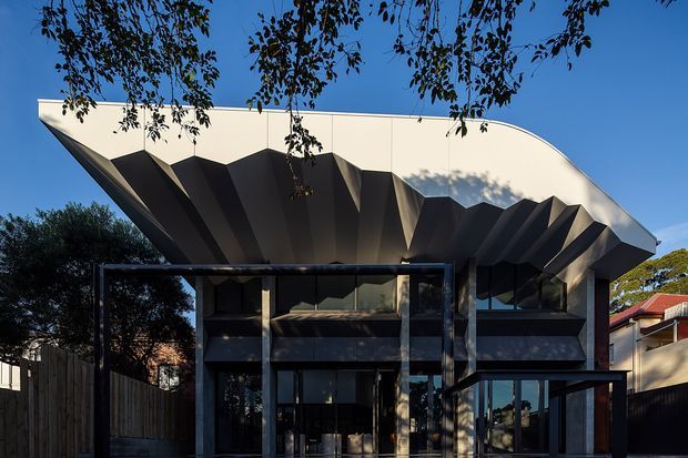 Exterior view of a striking contemporary multi-level building with a sculptural, dramatically folded overhanging roof or canopy. The dark glazed facade includes large windows and a glazed entrance, with residential buildings and vegetation surrounding the site.