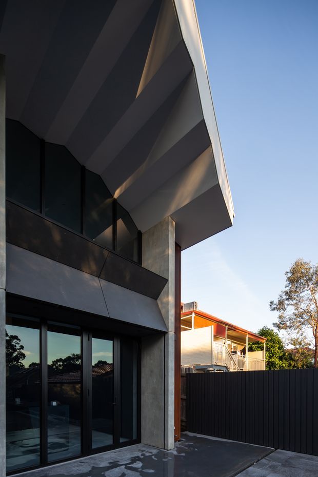 Photograph of a contemporary multi-level residential building exterior, focused on a dramatic cantilevered roof or upper-floor overhang. The facade combines dark metal cladding, large black-framed glazed doors and windows, exposed concrete, and a substantial concrete column. A paved terrace or driveway occupies the foreground, with neighbouring residential buildings and trees visible beyond a dark privacy fence.