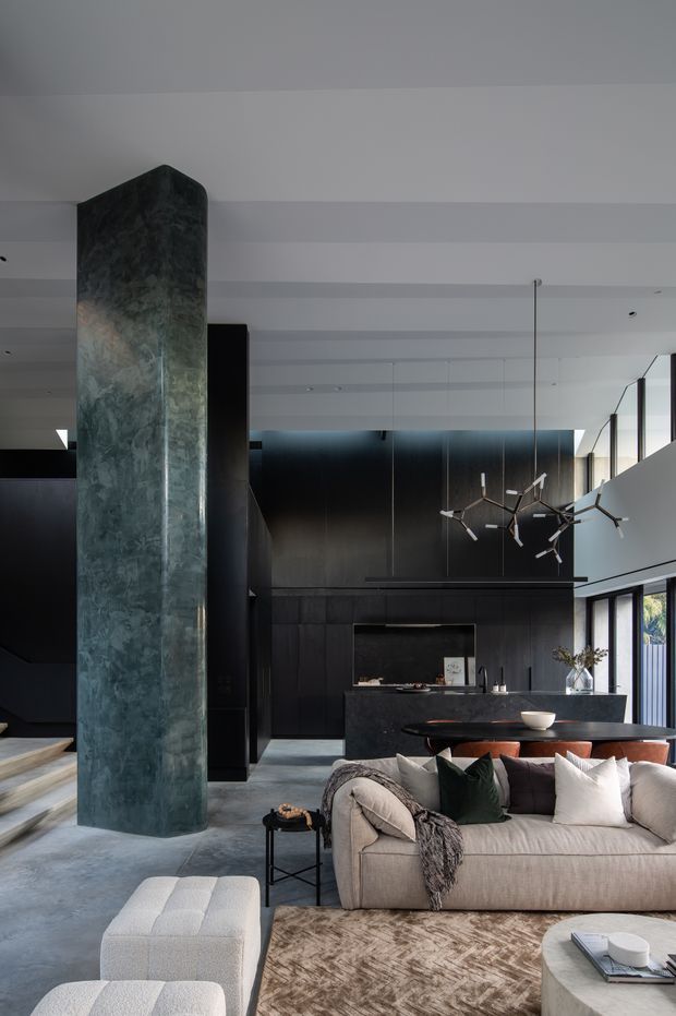 A sophisticated open-plan living room with a pale upholstered sectional sofa, textured rug, and low coffee table in the foreground. The space features dark built-in cabinetry, a linear fireplace, a stone kitchen island, dramatic pendant lighting, full-height glazing, and a tall sculptural dark stone-clad column. The palette is monochromatic and moody, balanced by warm timber accents and soft furnishings.