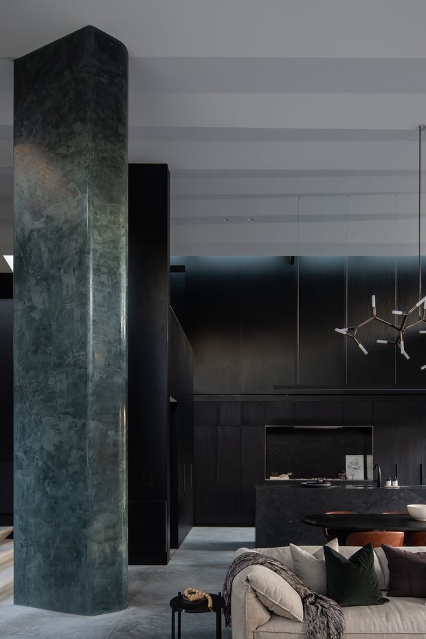 A dark, sophisticated open-plan interior showing a black kitchen with integrated cabinetry and island, a sculptural branching pendant light, and part of a living area with a large upholstered sofa. A tall rounded structural or feature column clad in mottled dark green-grey stone dominates the foreground. The space has a high white ceiling with horizontal detailing, dark wall panelling, polished grey flooring, and a moody, luxurious atmosphere.