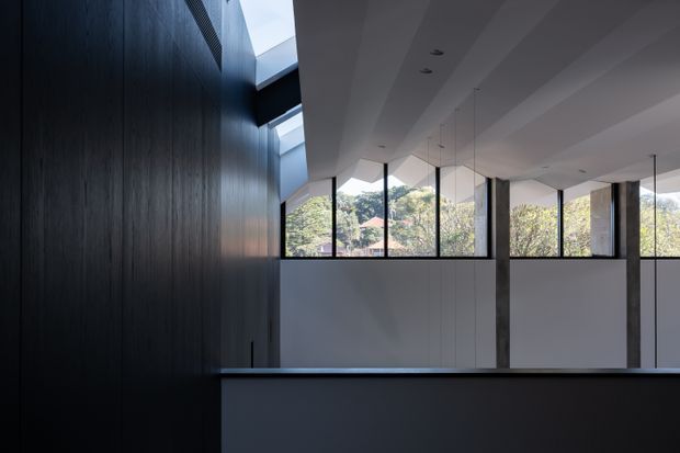 A contemporary interior with a dramatic dark timber feature wall, a long dark counter or balustrade in the foreground, and a bright upper-level space opening to extensive glazing. Slim black-framed windows, exposed concrete columns, recessed ceiling lights, and suspended linear pendant lights create a refined, minimalist architectural character. Trees and neighbouring roofs are visible outside through the windows.