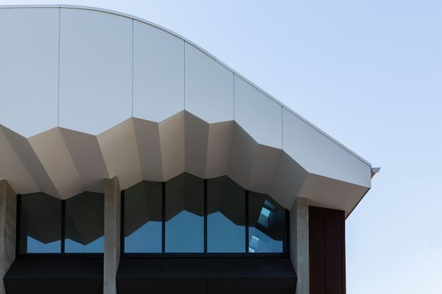 Close-up exterior view of a highly contemporary building facade, featuring a sweeping curved roofline, faceted folded soffit, dark full-height glazing, concrete structural elements, and a deep timber-toned entry door.