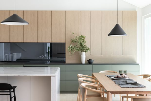 A refined open-plan kitchen and dining area featuring full-height light timber cabinetry, muted sage-green lower cabinets, a dark stone or glass splashback, and a pale stone island. A timber dining table with curved timber chairs sits beneath two black pendant lights. The space is bright, minimal, and softly contemporary.