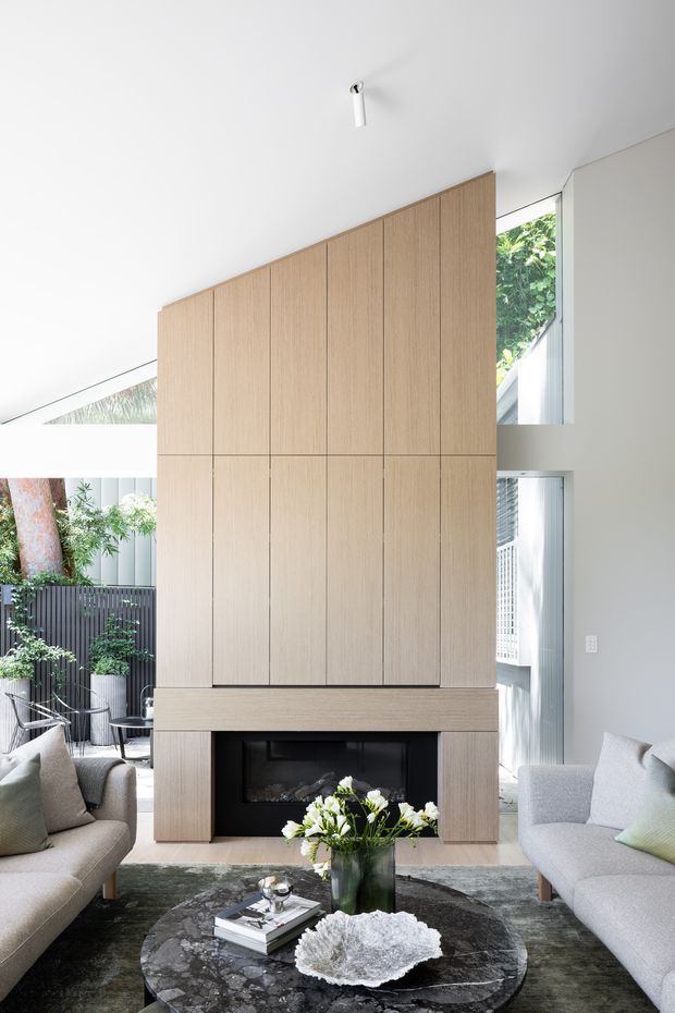 Bright contemporary living room centered on a tall fireplace wall clad in light timber-toned vertical panels. Two pale upholstered sofas face a dark round stone coffee table, with a floral arrangement and books. Large glazed openings connect the room to a leafy outdoor courtyard, while a high angled ceiling and abundant daylight create an airy, refined atmosphere.