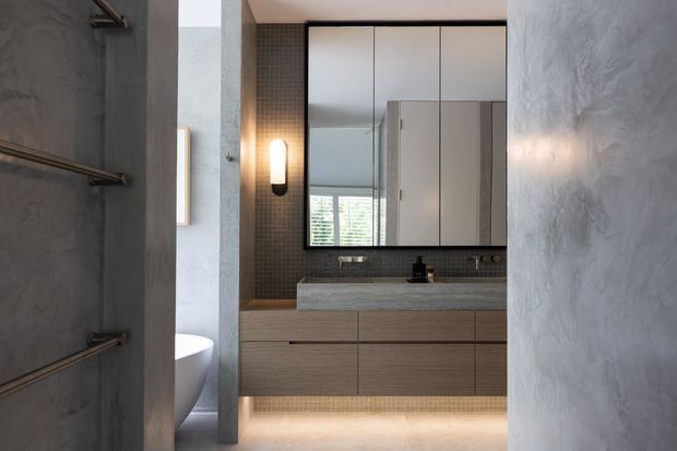 A contemporary bathroom viewed through a doorway, featuring a floating timber-look vanity with an integrated stone benchtop, dark small-format tiled splashback, large mirrored cabinetry, wall-mounted brass-toned towel rails, and concealed under-vanity lighting. A freestanding bathtub is partially visible at left. The room has textured grey walls and a refined, moody palette.