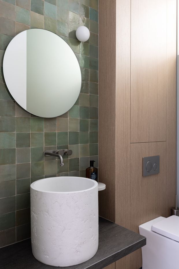 Contemporary bathroom featuring a round vessel basin on a dark benchtop, a large circular mirror, green-grey square tiled wall, brushed metal wall-mounted tapware, warm timber cabinetry or wall panelling, and a concealed cistern toilet. The space has a refined, earthy and softly lit material palette.