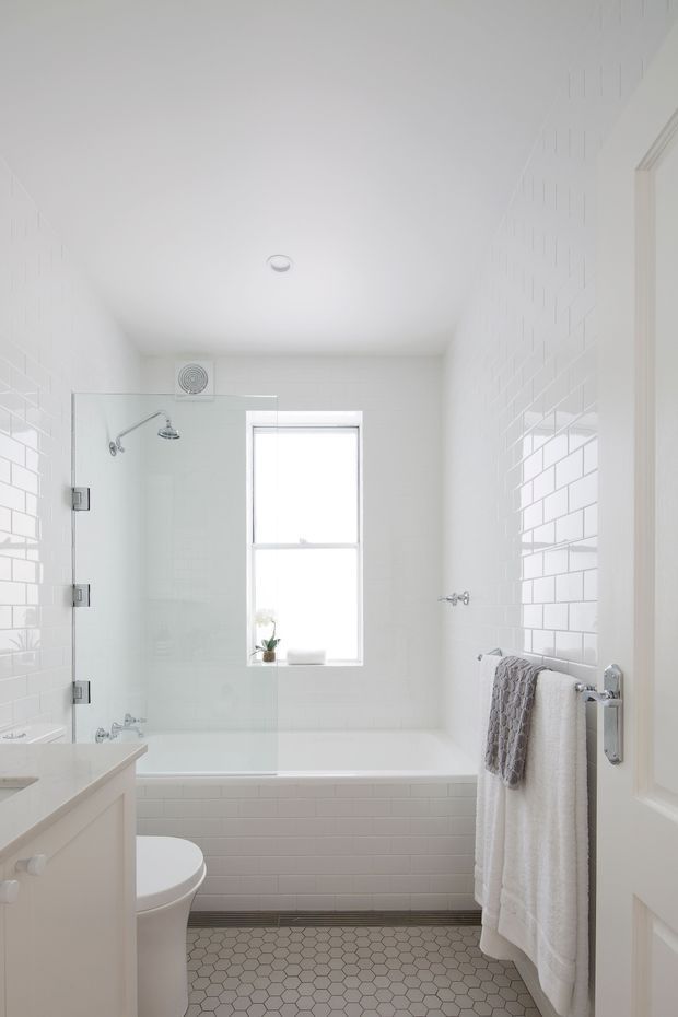 Bright, compact white bathroom with a bathtub and glass shower screen, white subway-tile walls, hexagonal tiled floor, pedestal-style toilet, vanity, central window, and chrome fixtures. Soft grey towels add a restrained accent.