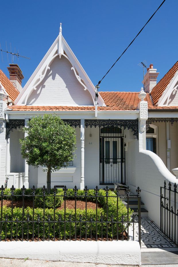 Street-facing view of a narrow heritage terrace or townhouse with a highly decorative white-painted facade. The building features ornate Gothic Revival gables, steep tiled roof sections, decorative timber bargeboards, cast-iron verandah lacework, a black security gate, patterned entry tiles, a small formal front garden, and black wrought-iron fencing.