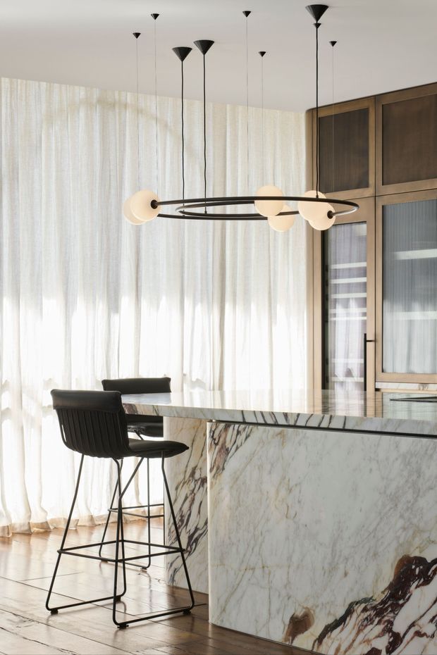 Bright, refined kitchen interior with a large veined marble island, dark bar stools, floor-to-ceiling sheer curtains, timber built-in cabinetry, and a sculptural black-and-opal globe pendant light. The space has a warm contemporary feel with abundant diffused daylight.