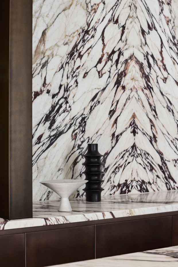 Close-up interior vignette featuring a dramatic bookmatched white marble feature wall and matching stone countertop over dark wood built-in cabinetry. A sculptural white bowl and black decorative vase sit on the surface, creating a refined, high-contrast composition.