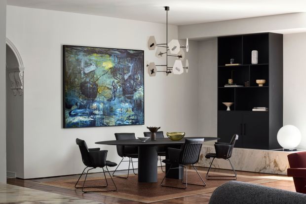 A refined dining area with a dark round dining table surrounded by contemporary black chairs, set against white walls and warm timber flooring. A large abstract blue and green artwork forms a feature wall, while a sculptural pendant light hangs above the table. Full-height black built-in cabinetry with open display shelving occupies the rear wall, with a pale stone-clad raised platform below. Arched decorative architectural detailing is visible at the left edge, and part of a living area appears in the foreground.