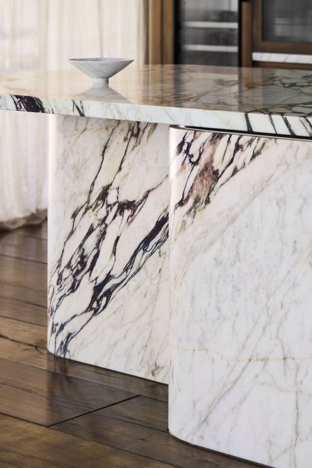 Close-up interior view of a sculptural marble kitchen island or counter, featuring dramatic dark veining across a white stone surface and waterfall-style curved cabinetry. The island sits on dark timber flooring, with sheer curtains and timber-framed glazing in the background. A small decorative bowl rests on the polished stone benchtop.