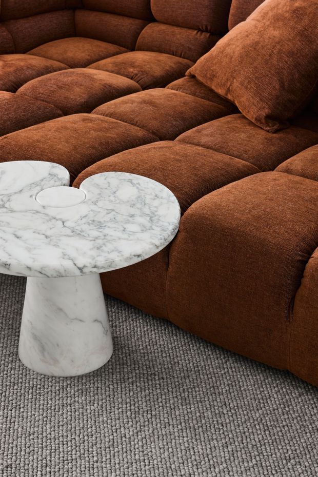Close-up interior detail of a large, plush, modular brown upholstered sectional sofa with deep tufted cushions, paired with a small white veined marble pedestal side table. A grey loop-pile carpet covers the floor. The composition is warm and tactile, focusing on furniture, upholstery, and material contrast rather than the wider room.
