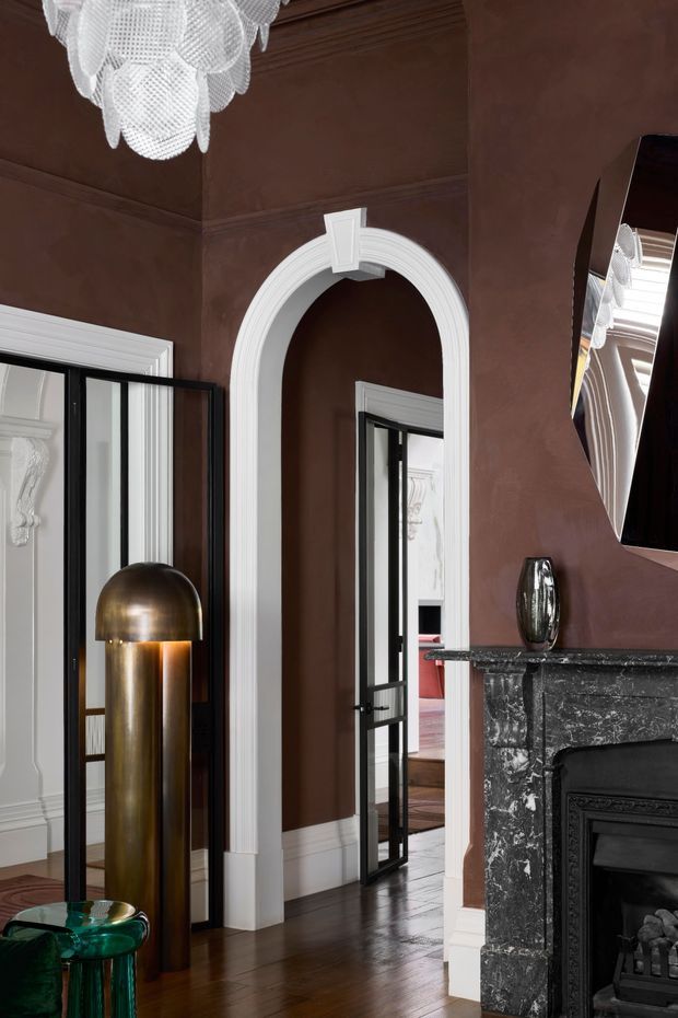 Elegant, richly styled interior corner with deep brown walls, ornate white arched and fluted door surrounds, dark timber flooring, black-framed glazed doors, and a substantial black marble fireplace with detailed surround. A sculptural brass floor lamp, crystal pendant light, abstract mirror, and green glass side table add dramatic decorative accents. The view appears to be from a living room or formal sitting area toward adjoining spaces.