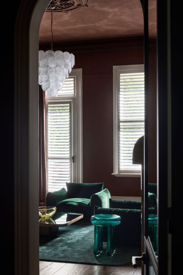 A richly styled sitting room viewed through an arched opening, featuring deep burgundy walls, tall shuttered windows, dark green velvet seating, a sculptural translucent teal side table, a patterned metallic ceiling, and a clustered white pendant light. A large mirror reflects the seating and adds depth. The room has a dramatic, moody, luxurious character with traditional architectural detailing and eclectic contemporary furnishings.